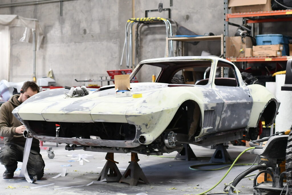 A C2 Corvette body stripped to primer on stands in the Nostalgia Hot Rods shop, with a builder working at the nose