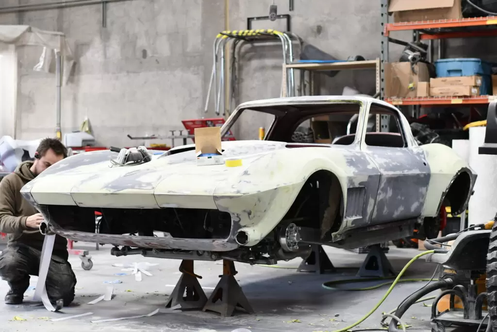 A C2 Corvette body stripped to primer on stands in the Nostalgia Hot Rods shop, with a builder working at the nose