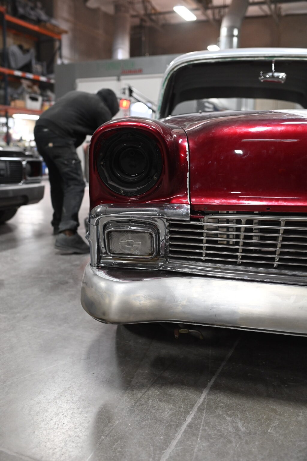The front end of a red Tri-Five Chevy on the Nostalgia Hot Rods shop floor, with a builder walking past
